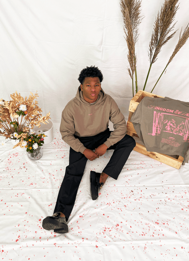 Model in olive green oversized hoodie with KINGDOM OF HEAVEN graphic, seated on a white backdrop with flowers and a t-shirt.